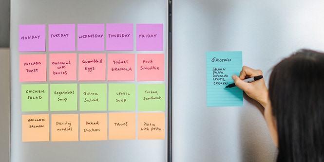 Un frigorífico con notas Post-it® que muestran un plan de comidas semanal, y una persona escribiendo una lista de la compra en una Nota Post-it&reg; con líneas
