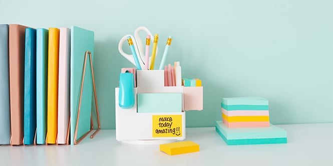 Un escritorio con cuadernos de colores, lápices, tijeras y notas Post-it®, creando un espacio de trabajo organizado y acogedor.
