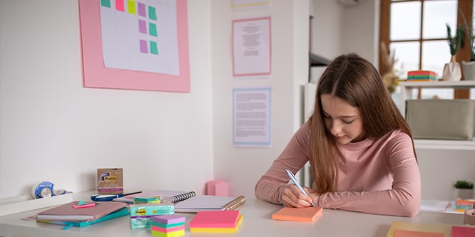 Estudiante anotando en notas Post-it® en un espacio de estudio con varios artículos de papelería, incluidas notas Post-it® y cuadernos
