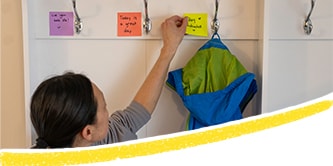 Mamá colocando Post-it® de colores con mensajes positivos y poniéndolos junto a perchas en el pasillo.
